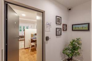 Un pasillo con una puerta que da a una habitación con una cama, un escritorio y una silla. Hay cuadros enmarcados en la pared y una planta en maceta.
