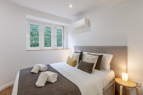 Una cama bien hecha con edredón gris y blanco, dos toallas enrolladas y una lámpara de noche.