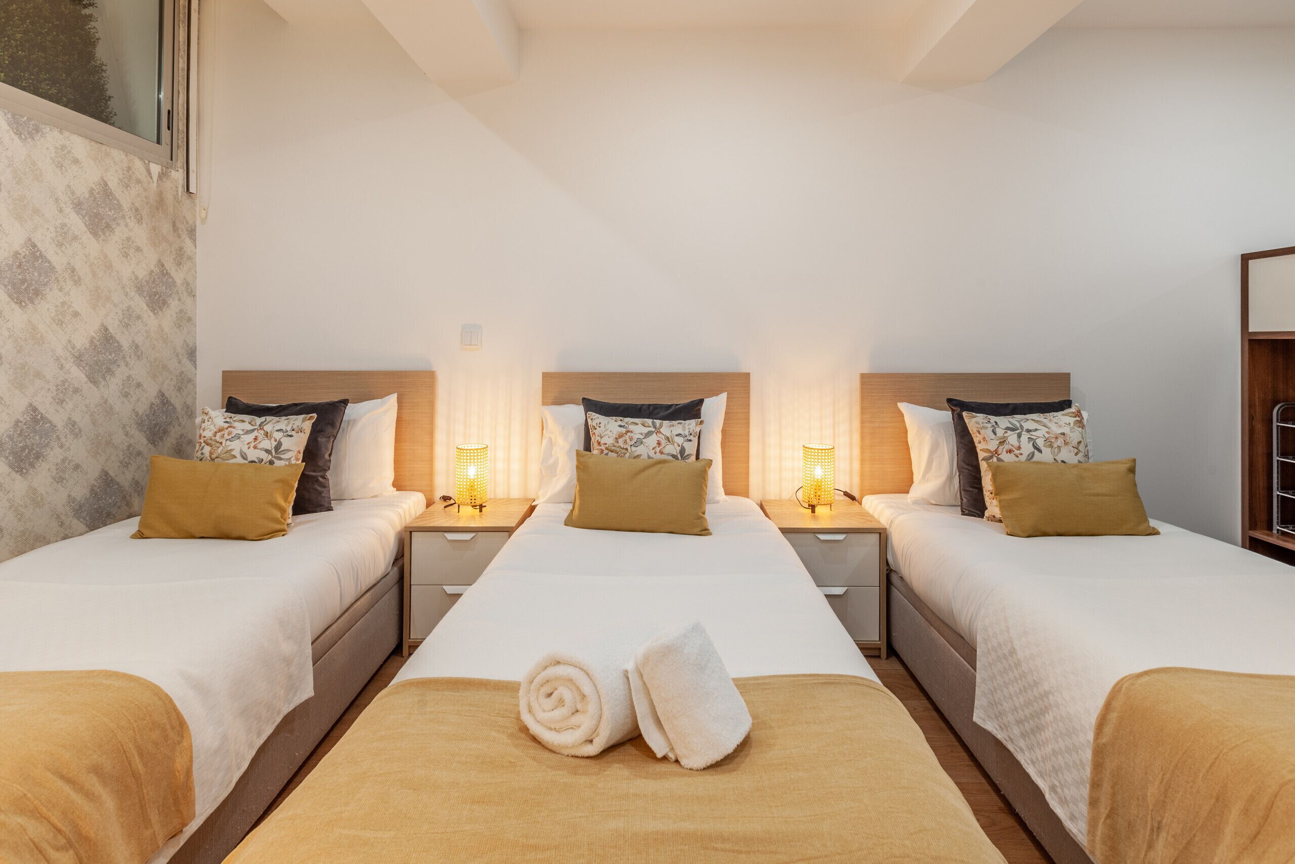 A hotel room with three beds, each with a wooden headboard, white bedding, and a brown throw blanket. There are two bedside tables with lamps, and a window with a view of trees.