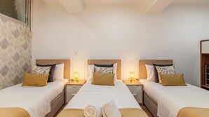 A hotel room with three beds, each with a wooden headboard, white bedding, and a brown throw blanket. There are two bedside tables with lamps, and a window with a view of trees. - Underground Batalha Studios - D (Oporto)