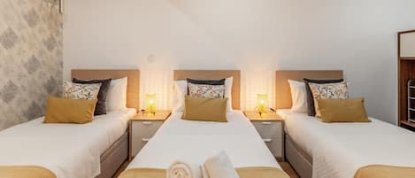A hotel room with three beds, each with a wooden headboard, white bedding, and a brown throw blanket. There are two bedside tables with lamps, and a window with a view of trees.