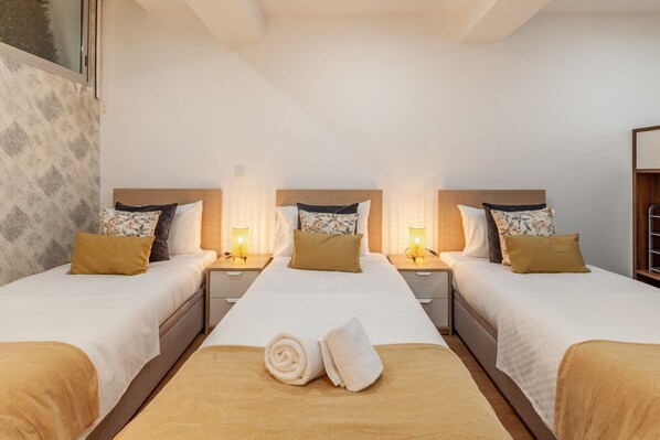 A hotel room with three beds, each with a wooden headboard, white bedding, and a brown throw blanket. There are two bedside tables with lamps, and a window with a view of trees.