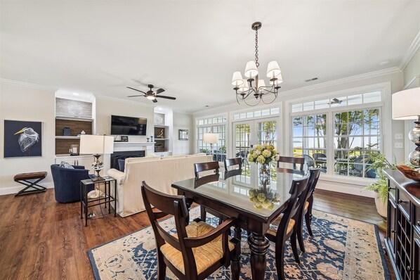A spacious dining area with a large table, chairs, and a chandelier. There is a ceiling fan and a television in the background.