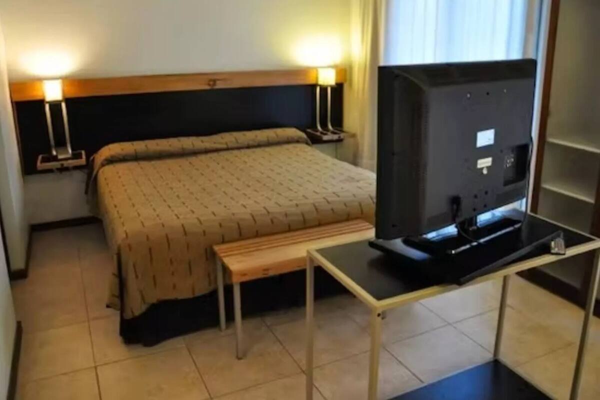 A hotel room with a bed, bedside lamps, a television on a stand, and a small table.