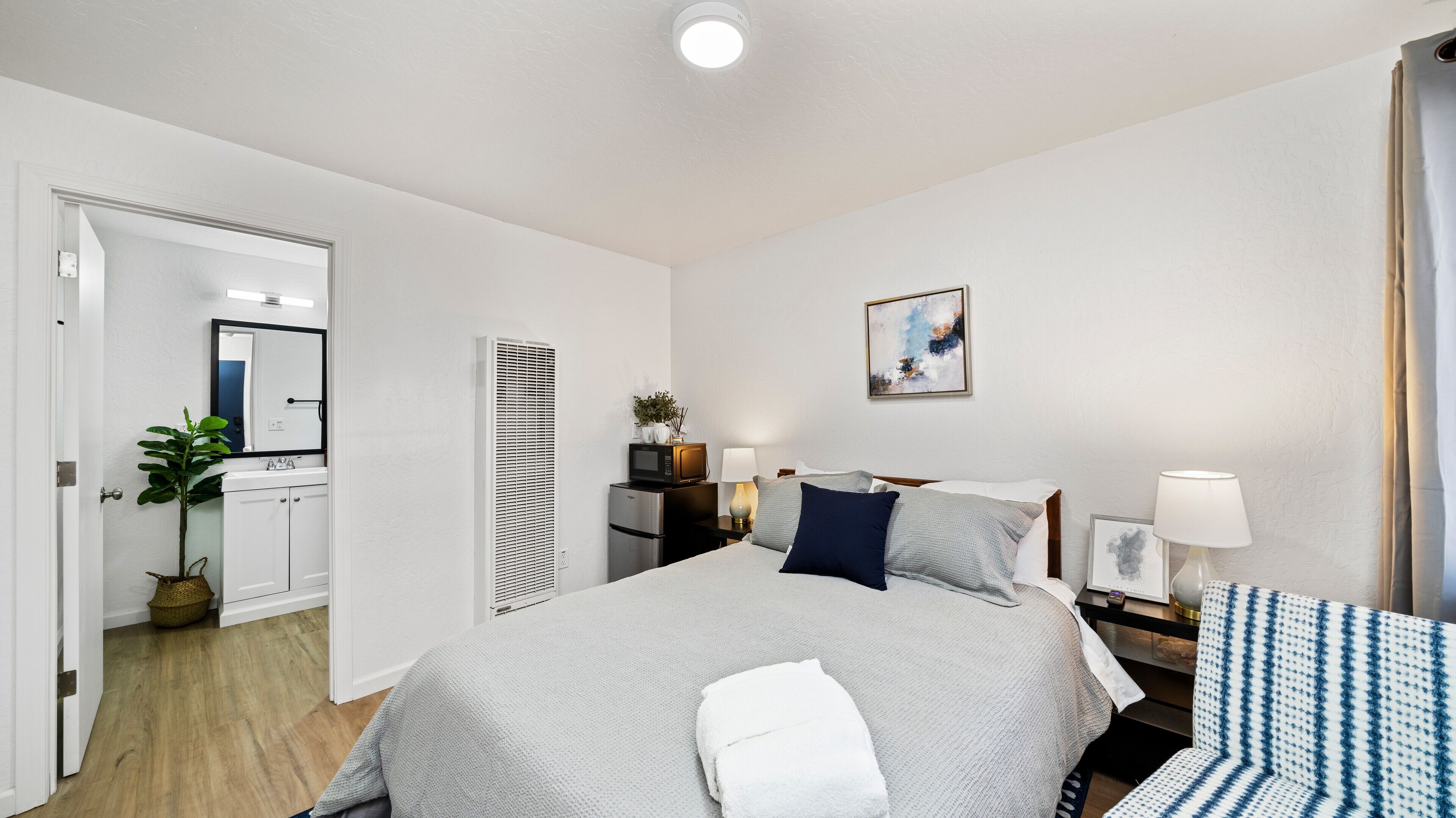 Une chambre d’hôtel avec un lit, des tables de chevet, une lampe, un tableau et une salle de bain visible à travers une porte ouverte.