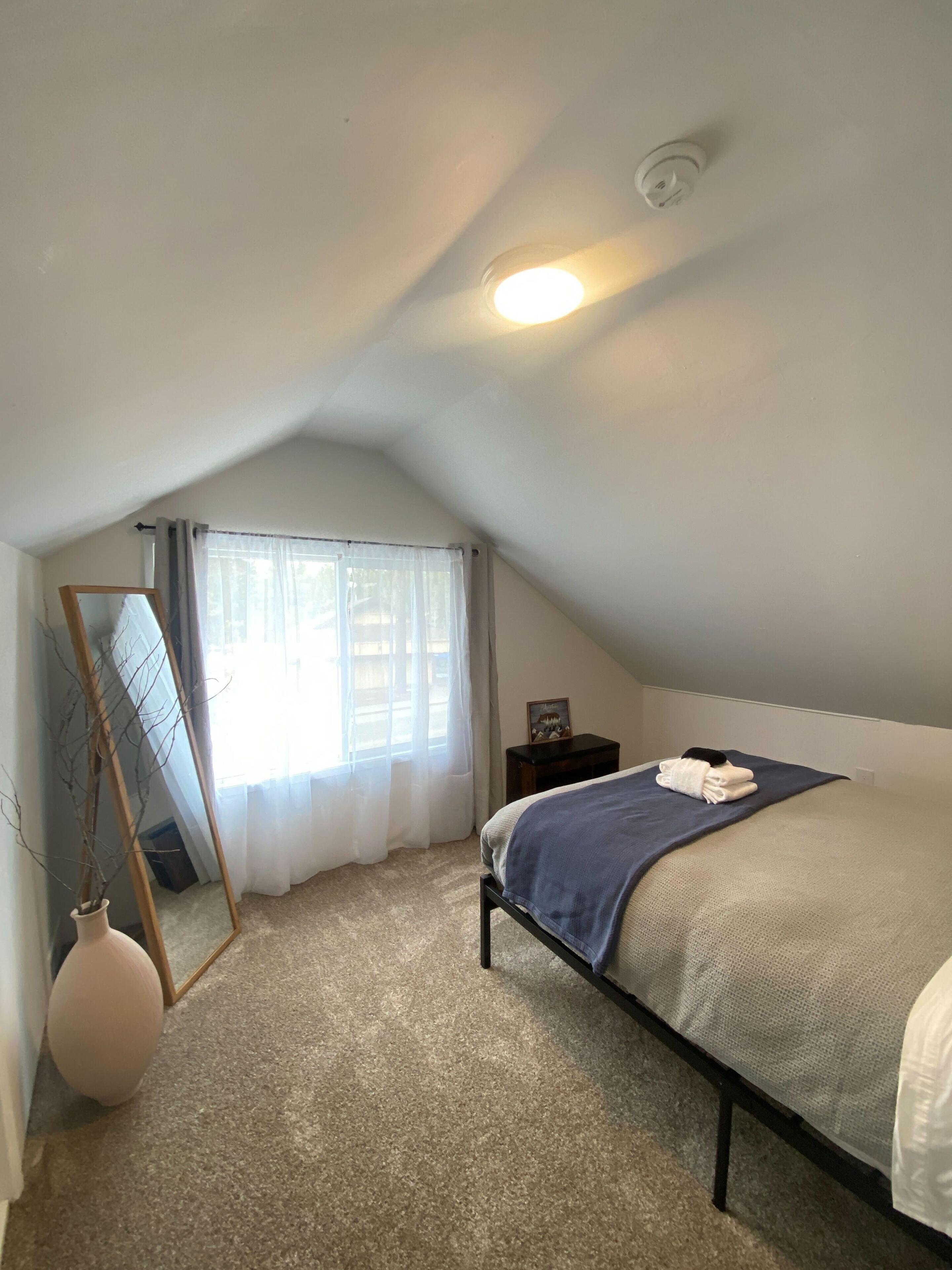 A bedroom with a bed, a bedside table, a mirror, a window with curtains, and a ceiling light.