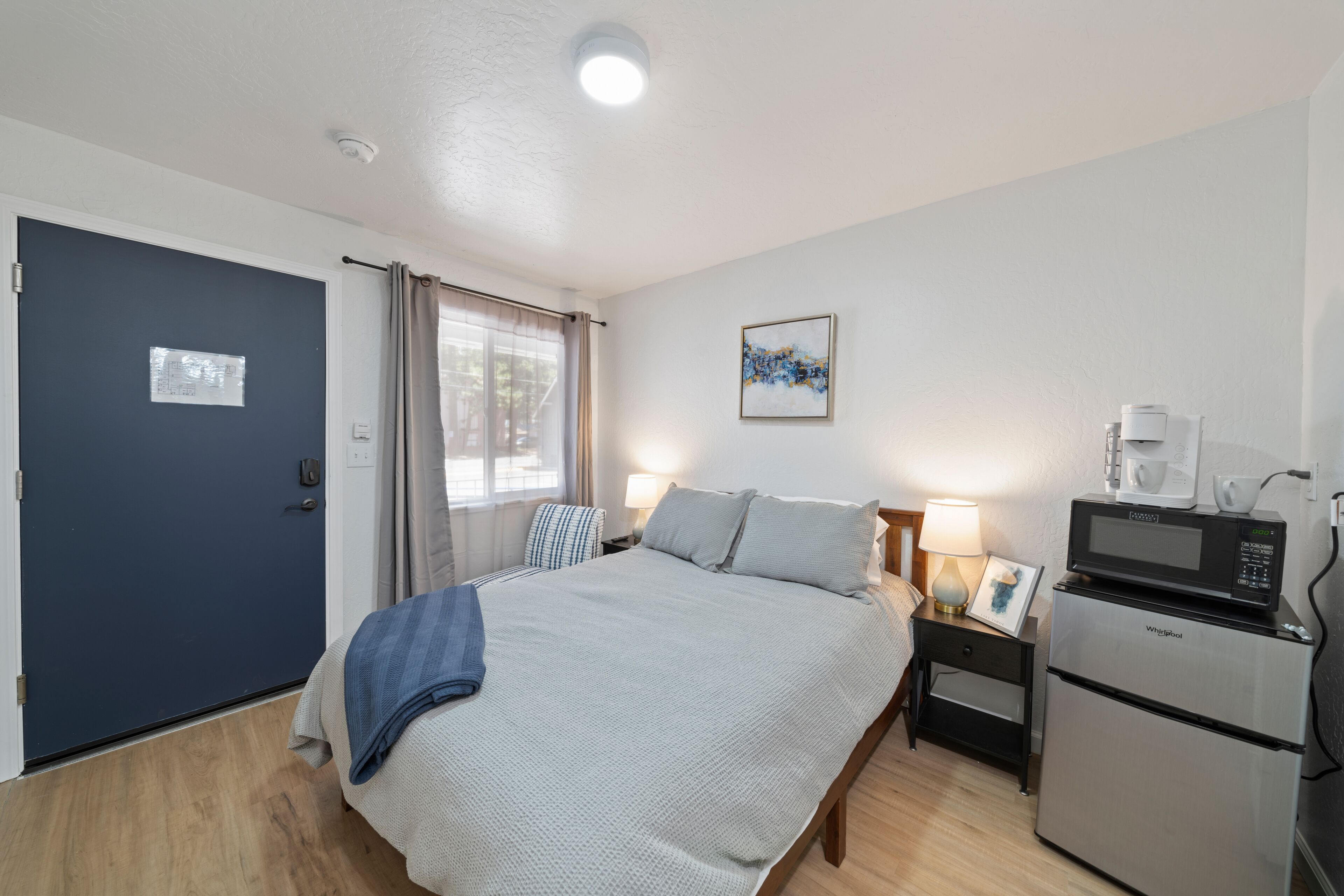 A hotel room with a bed, a desk with a lamp, a microwave, and a refrigerator.