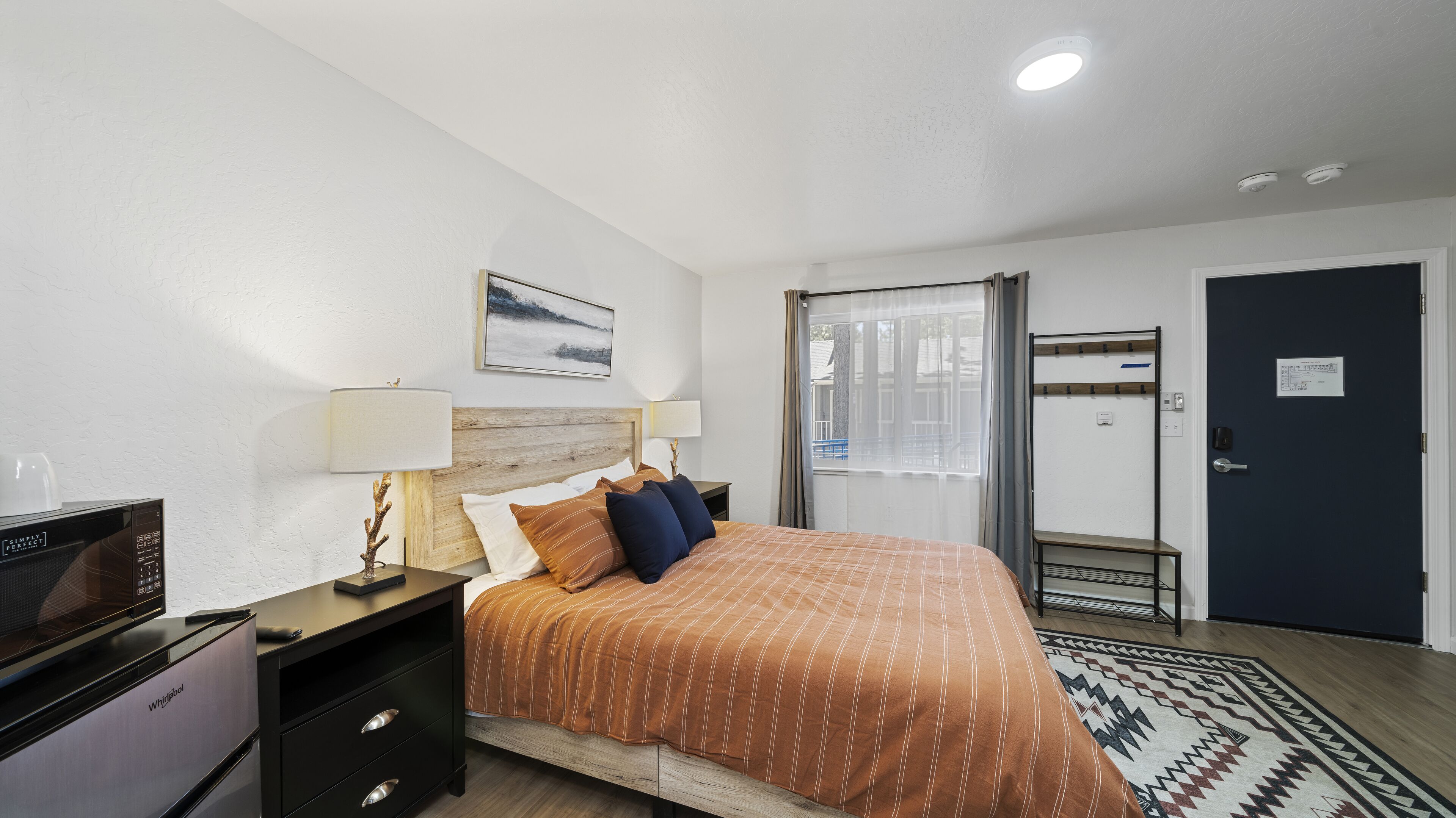 A hotel room with a bed, a desk with a microwave, a lamp, a picture on the wall, a window with curtains, and a door with a coat rack.