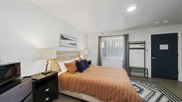 A hotel room with a bed, a desk with a microwave, a lamp, a picture on the wall, a window with curtains, and a door with a coat rack.