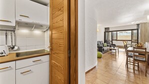 Una cocina moderna con armarios blancos y tiradores de madera, un comedor con mesa y sillas, y una sala de estar con sofá y ventana.