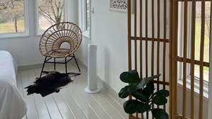 Una habitación con cama, una silla de mimbre, una planta en maceta, un adorno en la pared y una ventana con vista a árboles.