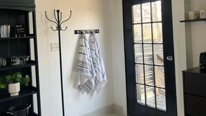 Una habitación con techo de madera, una puerta negra con ventana, un perchero con toallas, un estante negro con utensilios de cocina y una alfombra de baño con un mensaje.