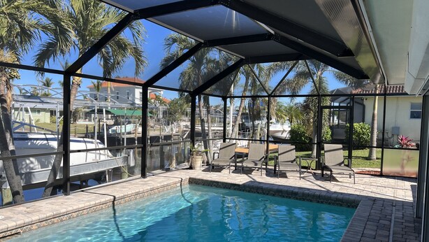 A covered swimming pool with clear blue water, surrounded by lounge chairs and a view of boats and palm trees. - Villa Nautica - Perfect for boaters (Cape Coral)