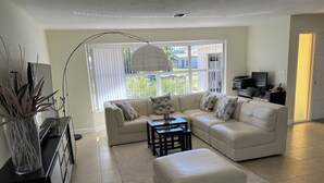 A modern living room with a beige sofa, a coffee table, and a TV. There is a large window with blinds, a plant, and a door leading outside. - Villa Nautica - Perfect for boaters (Cape Coral)