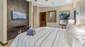 A bedroom with a large bed, a flat-screen TV mounted on a brick wall, a ceiling fan, and a window with a view of the outdoors. - Slippery Rock Vacation Home - sleeps 9 (Flagstaff)