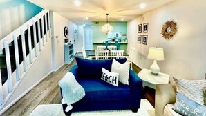 A living room with a blue sofa, a white rug, and a small table with a lamp. There is a staircase on the left and a kitchen area in the background.