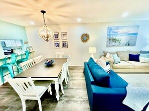A living room with a blue sofa, a dining table with chairs, a kitchen area, and a wall with framed pictures and a decorative wreath. - Manatee Beach Beauty! 1 minute walk to beach, restaurants and shops 🏖️ (Holmes Beach)
