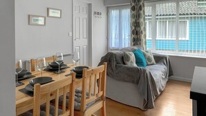A cozy living room with a dining area, a sofa with cushions, and a window with curtains. - A lovely contemporary getaway, just a few steps away from the beautiful beach. (Mundesley)