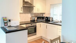A compact kitchen with white cabinets, a black countertop, and a built-in oven. There is a dining table with chairs and a coffee maker. - A lovely contemporary getaway, just a few steps away from the beautiful beach. (Mundesley)