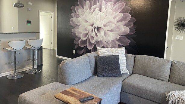 En moderne stue med en grå modulsofa, et træbord med en fjernbetjening, og et stort blomster-vægmaleri.