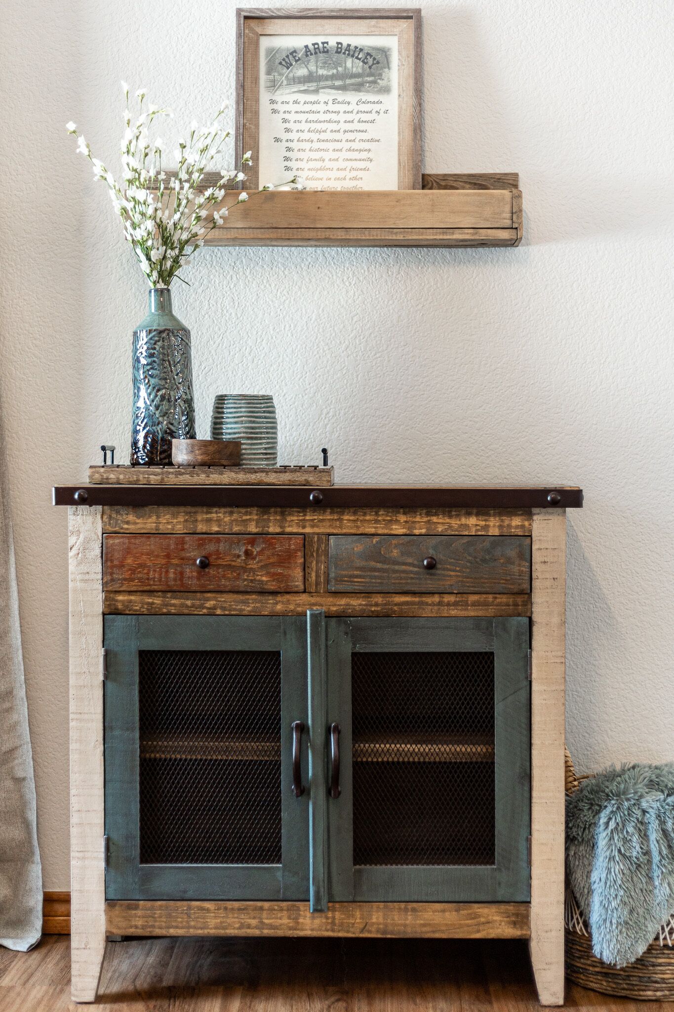 A rustic wooden cabinet with two doors, a vase with flowers, and a framed quote on the wall above.