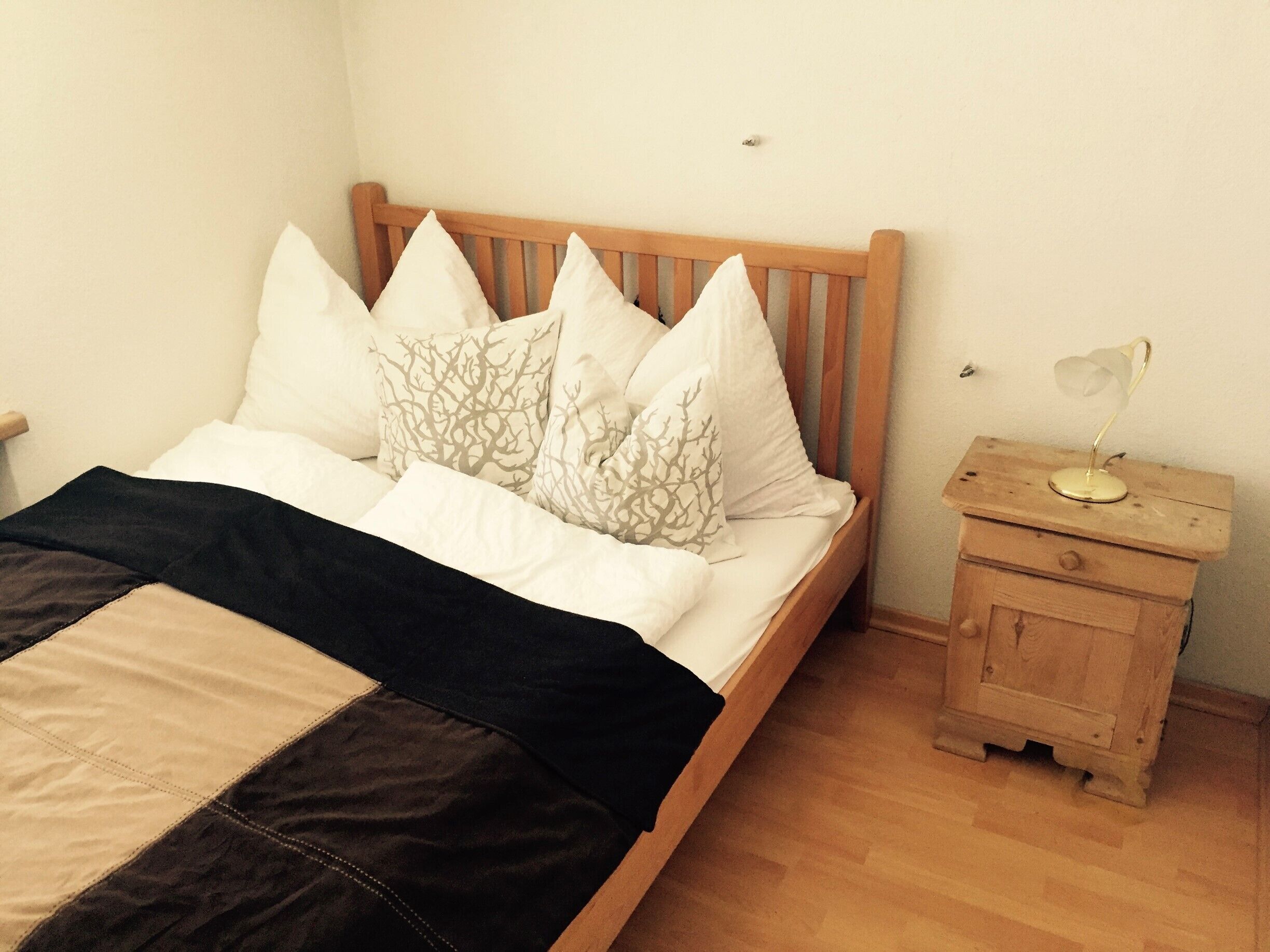 Een net opgemaakt bed met een houten hoofdbord, een nachtkastje met een lamp en een klein houten kastje.