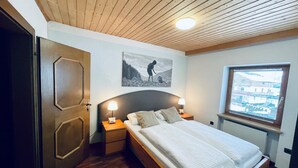 A bedroom with a wooden ceiling, a bed with white linens, two bedside tables with lamps, a window with blinds, and a door. - Gemütliche Ferienwohnung im Dorfzentrum mit Garten für 2- 6 Personen (Aschau im Zillertal)