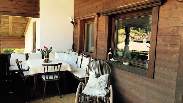 A wooden cabin with a dining area featuring a table and chairs, a wicker chair, and a large window with a view of a house and greenery. - Gemütliche Ferienwohnung im Dorfzentrum mit Garten für 2- 6 Personen (Aschau im Zillertal)