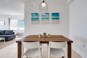 Uma área de refeições com mesa de madeira, cadeiras brancas e três obras de arte com tema praiano.