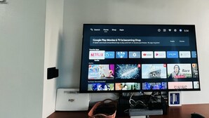 Un televisore a schermo piatto montato a parete, con uno scaffale sottostante dove si trovano un dispositivo di streaming e un telecomando.