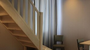 Ein Zimmer mit einer Holztreppe, einem Tisch, zwei Stühlen und einem Fenster mit Vorhängen.