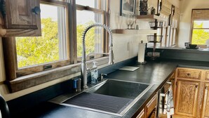 A kitchen with wooden cabinets, a black countertop, a stainless steel sink, and a window with blinds. - Three-bedroom house with views, decks, golf cart and home gym! (Ocracoke)