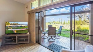 Un salon moderne avec un téléviseur posé sur un meuble en bois, une porte-fenêtre coulissante donnant sur une terrasse extérieure avec une table et des chaises, et une grande fenêtre offrant une vue sur l’extérieur.