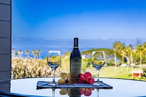 Une table sur laquelle se trouvent une bouteille de vin Caymus, deux verres de vin et deux fleurs, le tout posé sur une table en verre offrant une vue sur l’océan et les palmiers.
