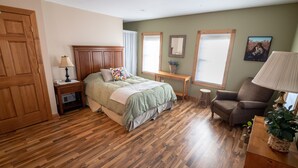 Une chambre avec un plancher en bois, un lit recouvert d’une couverture verte, une table de chevet avec une lampe, une chaise et une fenêtre donnant sur un beau paysage.