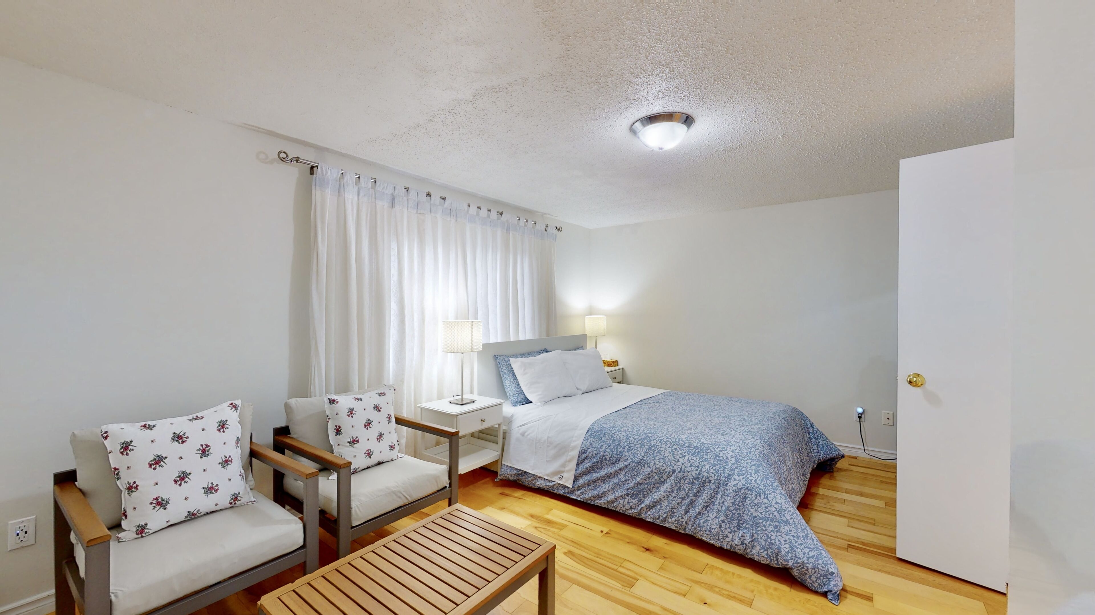 A small, clean room with a bed, two chairs, a nightstand, and a wooden table.