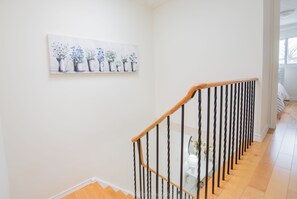 Una escalera con barandilla de madera y barandillas de metal negro, que conduce a una habitación con pared blanca y un cuadro de plantas en macetas.