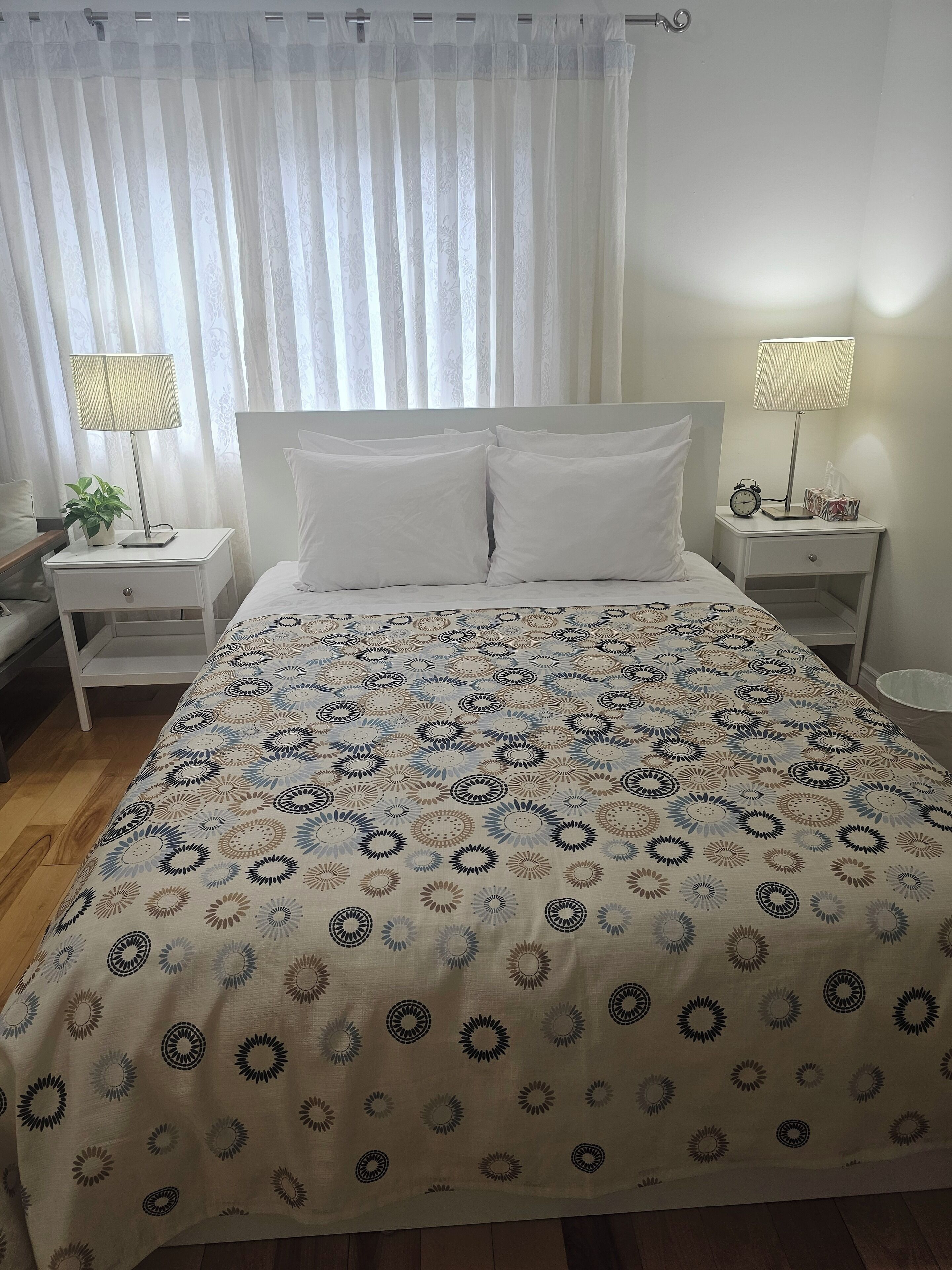 A neatly made bed with a patterned bedspread, two bedside tables with lamps, a clock, and a small plant on one of the tables.
