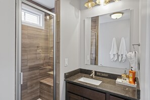 A modern bathroom with a glass shower enclosure, a large mirror, and a dark countertop with a sink and toiletries. - On The Rocks (Ottawa)