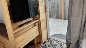 Ein Fernseher mit Flachbildschirm steht auf einem Holztisch, zusammen mit einer Fernbedienung. Eine Schiebetür aus Glas führt auf eine Holzterrasse, und auf dem Boden liegt ein gemustertes Teppich.