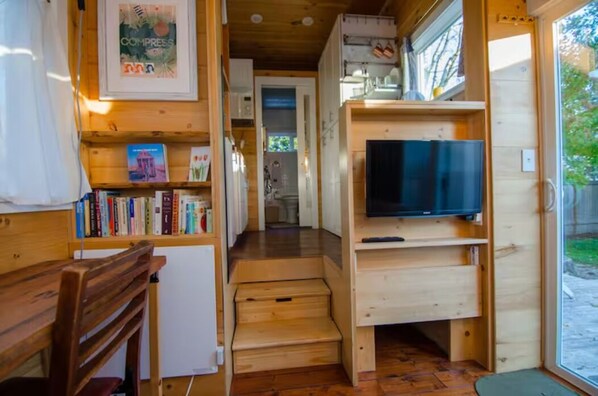 A compact room with a wooden bench, a bookshelf with books, a television on a wooden stand, and a door leading to another room. - Tacoma Tiny House (Tacoma)