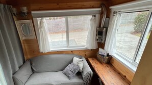 Ein gemütliches Blockhaus-Interieur mit Holzbalkendecke, einem grauen Sofa mit Kissen, einem kleinen Tisch mit einem 'GUEST'-Schild und einem Fenster mit Vorhängen.