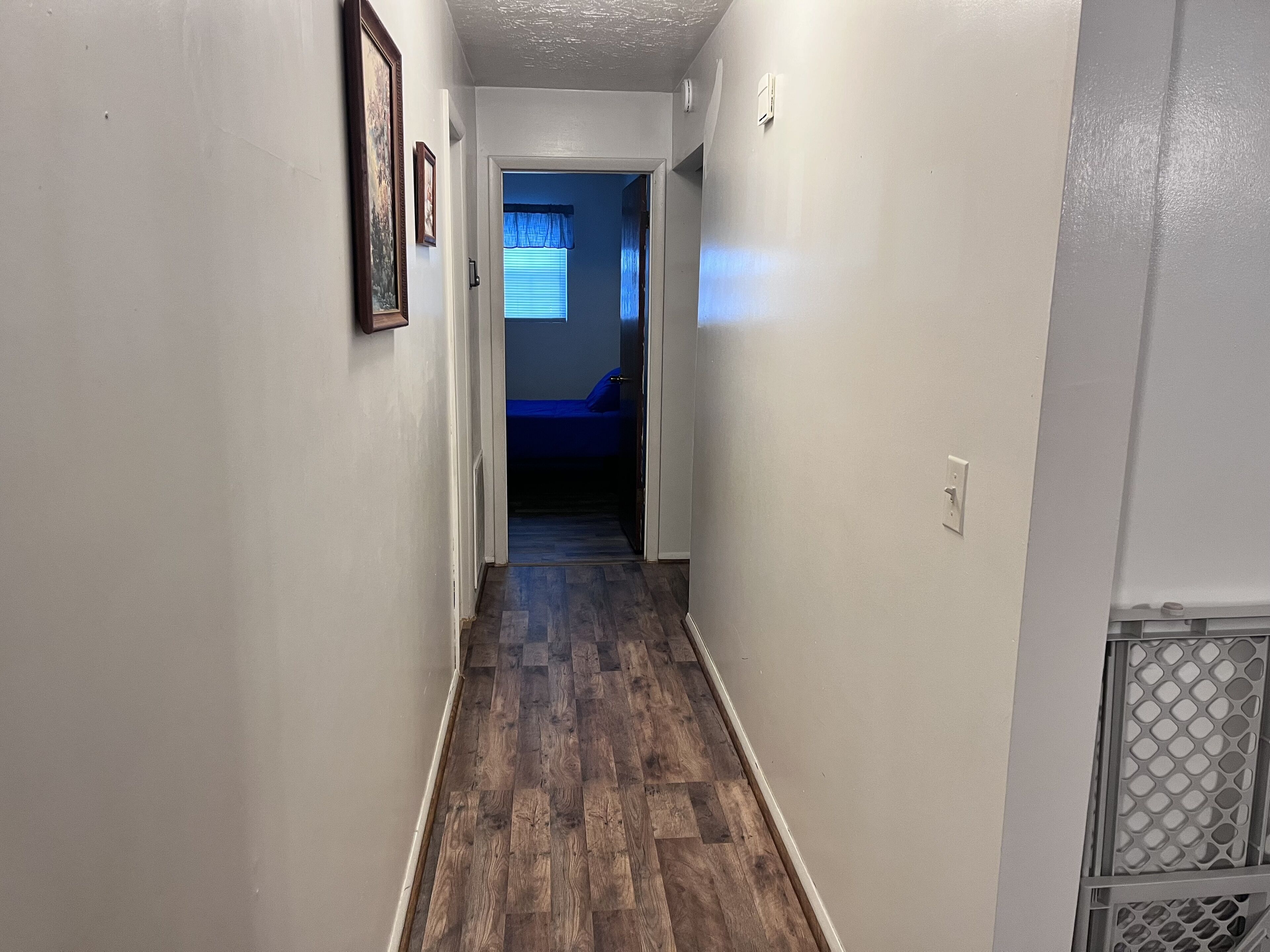 A narrow hallway with wooden flooring, white walls, and a door leading to a bedroom with a blue bedspread.