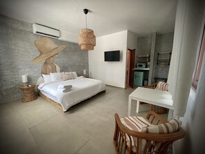 A modern bedroom with a large bed, a dining area with wicker furniture, a television, and a kitchenette. - Corcega Beachfront Suites   (Rincon)