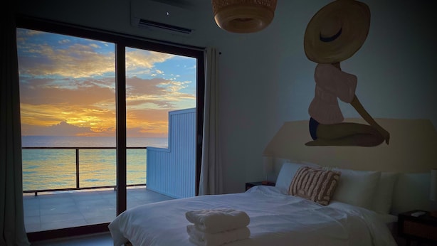 A bedroom with a bed, a nightstand, a lamp, and a view of the ocean at sunset.