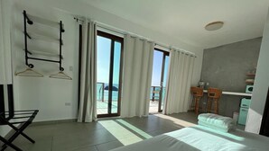 A hotel room with a large bed, a seating area with a table and chairs, a view of the sea through the windows, and a balcony with a chair.