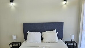 Ein ordentlich bezogenes Bett mit weißen Laken, zwei Nachttische mit Lampen sowie zwei Wandleuchten.