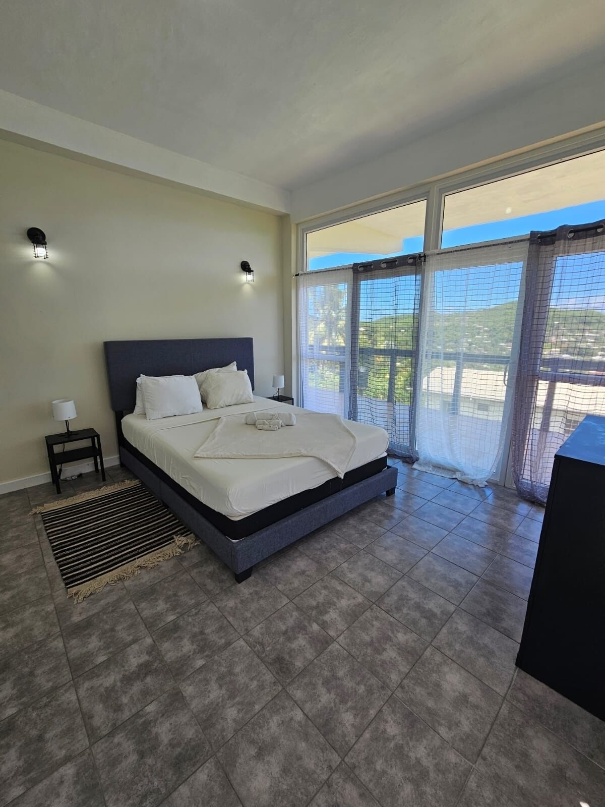 A bedroom with a large bed, two bedside tables, a rug, and a view of greenery through the window.