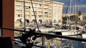 A marina with multiple boats docked, a building with balconies in the background, and a potted plant on a balcony railing.