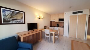 A compact living space with a sofa, a TV, a dining table with chairs, a kitchen area with a microwave, and a framed picture on the wall.